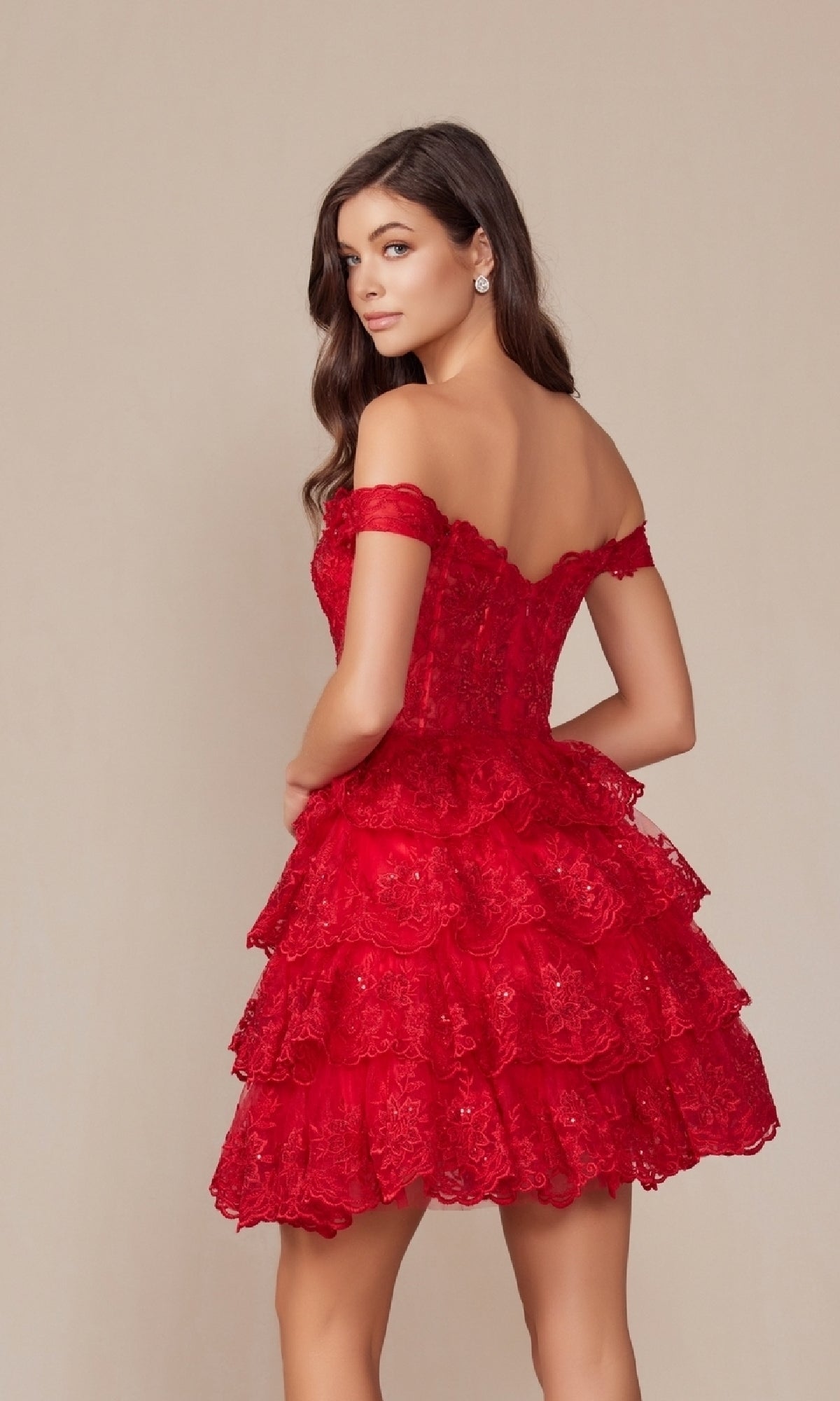 A woman models the Nox Anabel R877 Short Ruffled Lace Hoco Dress, featuring off-the-shoulder tiered ruffles, as she looks over her shoulder against a neutral background.